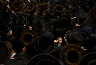A young Orthodox Jewish boy surrounded by hundreds of Orthodox Jewish men wearing black coats and hats. The men are gathered to see their spiritual leader who has arrived from Antwerp.