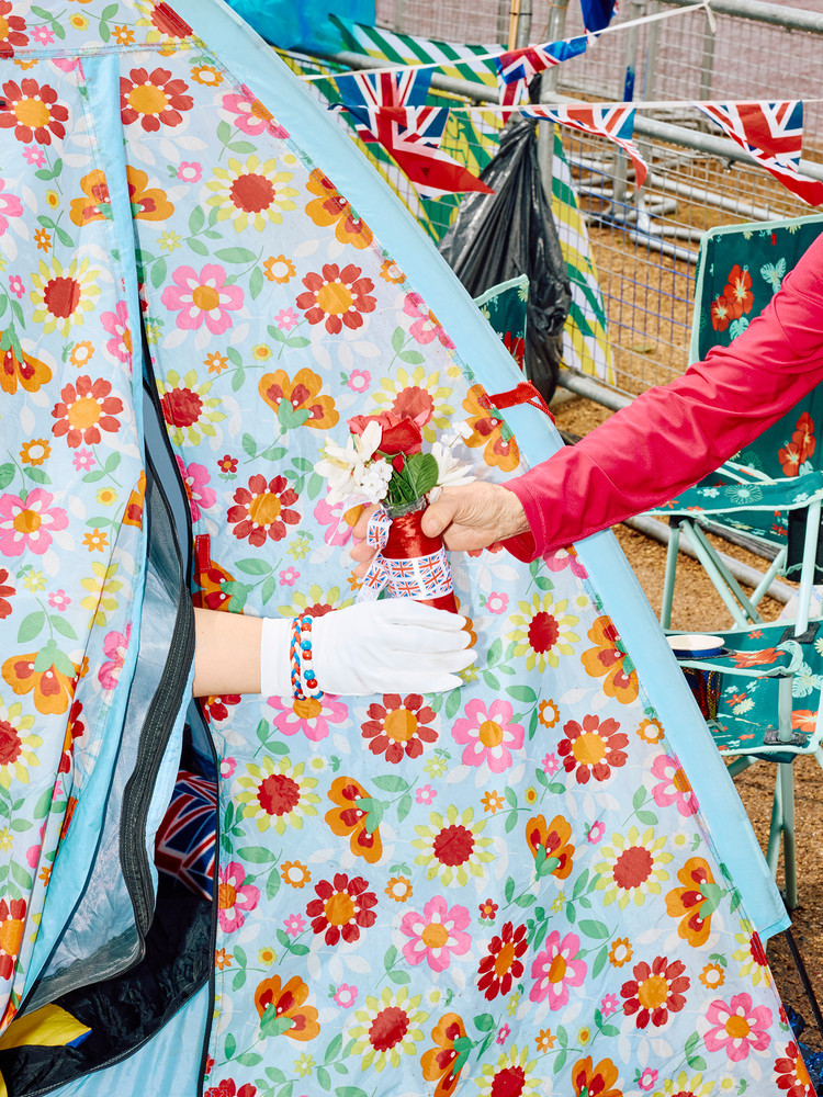 A royal fan wearing a white glove and Union Jack-themed bracelets hands over flowers to a friend from inside a tent erected along the Mall in London on May 5, 2023 on the eve of the coronation of Britain’s King Charles III. © Benjamin Cremel