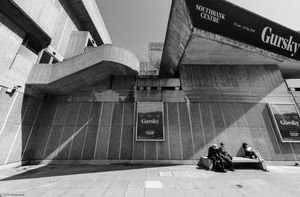 Tourists seated on a bench behind the Hayward Gallery on the South Bank, London | 01