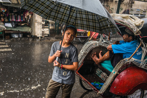 A child rents an umbrella from a local business and hopes for a tip from  costumers by sheltering from the heavy rains. Yogiakarta