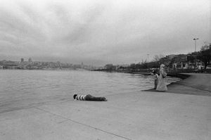 A boy looks in the water while his mother and brother stand nearby in a waterside park.