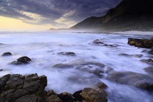 Motion / A seascape of the rugged coast on the Atlantic Ocean, along the Cape Peninsula, South Africa. A double long exposure with rocks and blurred waves in the foreground and mountains and cloudy sky at sunset in the background.